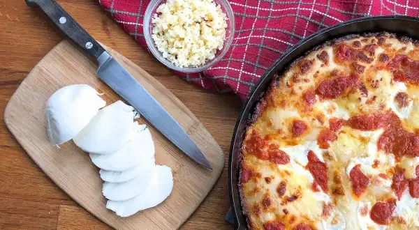 Idealny jaki ser do pizzy? Na desce świeża mozzarella, obok nóż i miseczka z tartym serem. Obok pyszna, gorąca pizza.