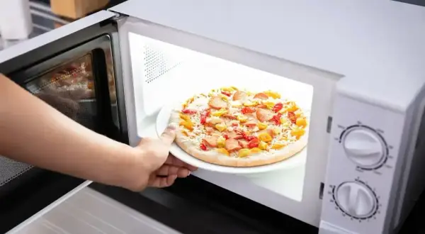 Ręka wkłada talerz z mrożoną pizzą do mikrofali. Szybki obiad gotowy!