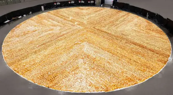 Ogromna, okrągła pizza, największa na świecie, rozciąga się na dużej przestrzeni. Jej powierzchnia pokryta jest równomiernie serem i przyprawami, tworząc wzór przypominający krzyż.