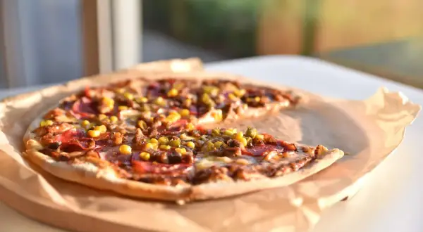 Pyszna pizza z kukurydzą, salami i oregano, podana na papierze do pieczenia, w słonecznym świetle.