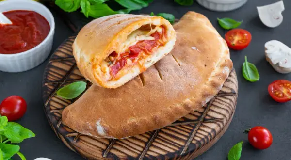Złocisty calzone, przekrojony na pół, odsłaniający gorące nadzienie z pomidorów i sera. Obok sos pomidorowy i świeża bazylia.