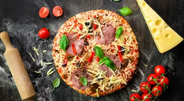 Gotowa pizza z salami i serem, obok wałek, pomidorki i ser. Idealna do domowego wypieku, zastanawiasz się, jaka mąka do pizzy domowej będzie najlepsza?