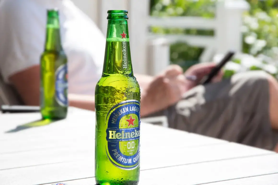 Orzeźwiająca butelka Heineken 0.0 procent, idealna na letni relaks. W tle osoba korzystająca z telefonu.