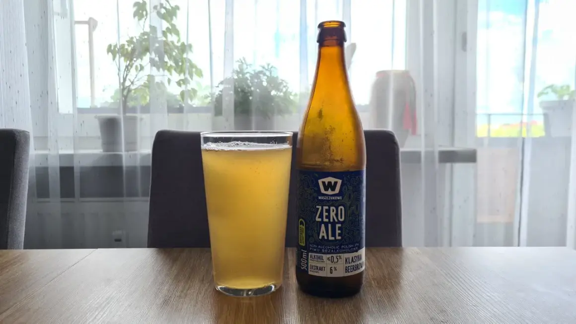 Szklanka i butelka piwa zero na drewnianym stole. Za nimi okno z firanką i roślinami.