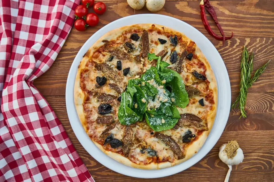 Idealna pizza z piwem na drewnianym stole, ozdobiona rukolą i parmezanem. Obok czosnek, pomidorki i rozmaryn.