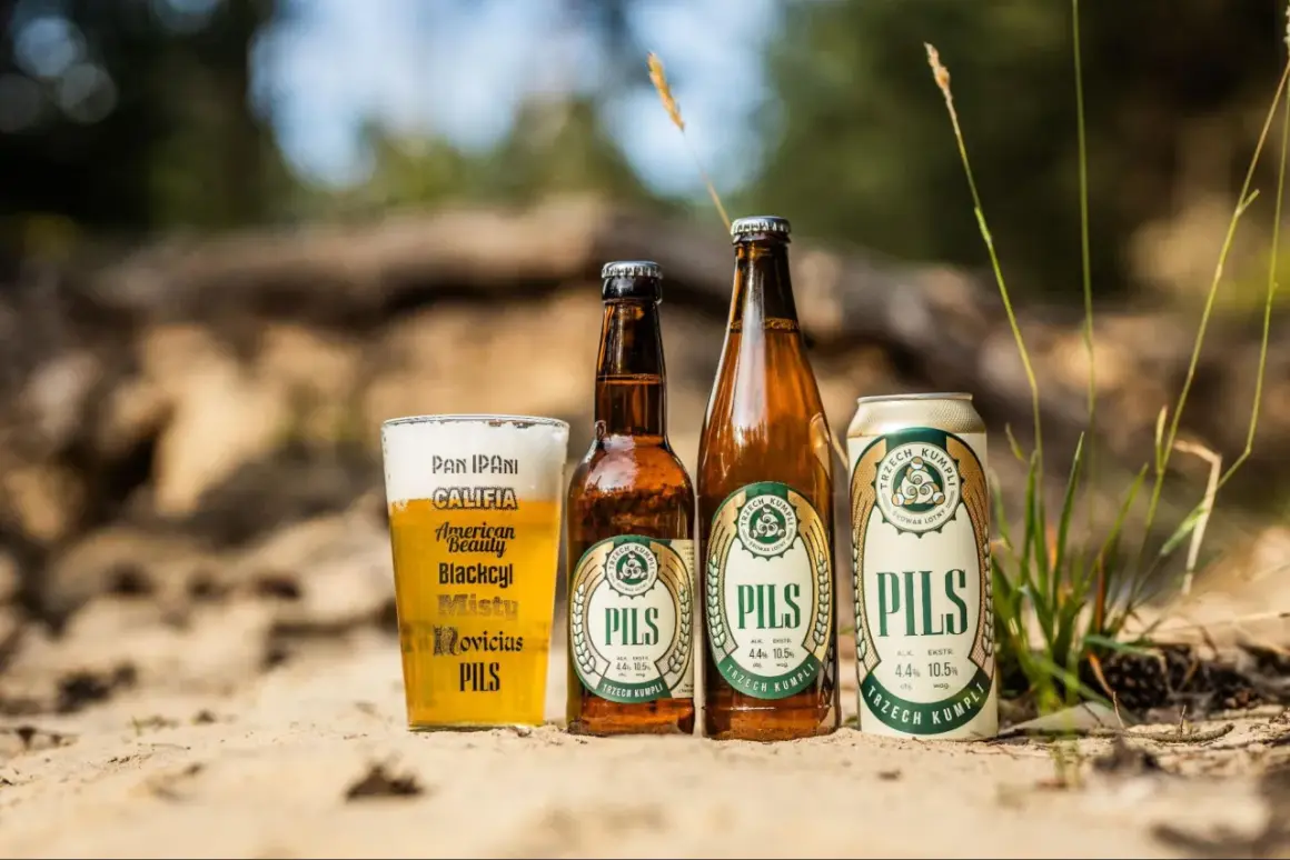 Szklanka, butelka i puszka piwa lager "Pils" od Trzech Kumpli na piasku, wśród traw.