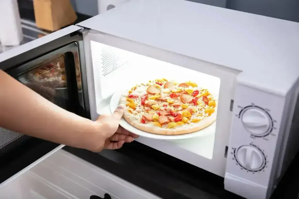 Ręka wkłada do mikrofali talerz z pizzą Giusseppe. Szybkie danie gotowe do podgrzania.