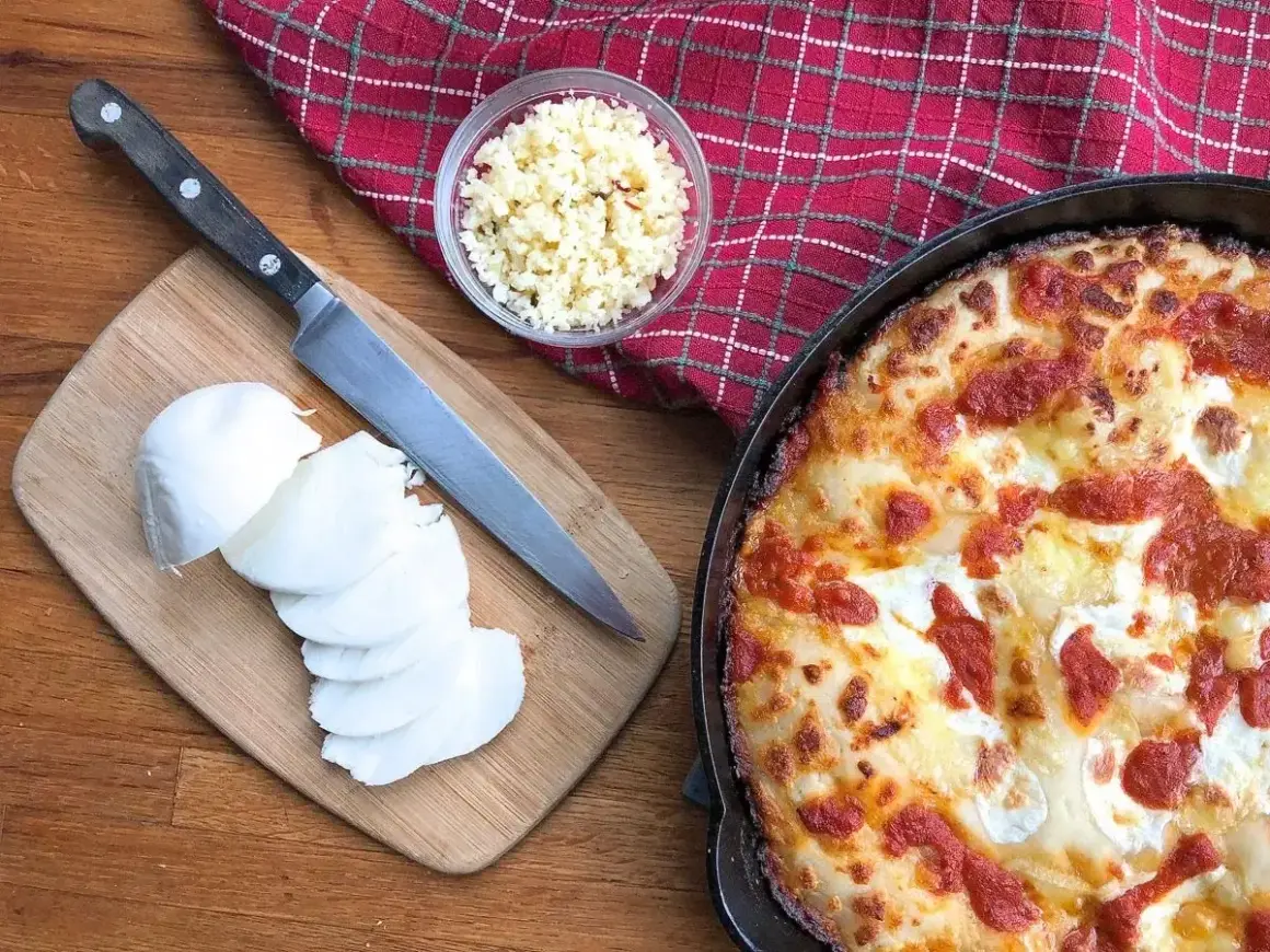 Idealny jaki ser do pizzy? Na desce świeża mozzarella, obok nóż i miseczka z tartym serem. Obok pyszna, gorąca pizza.