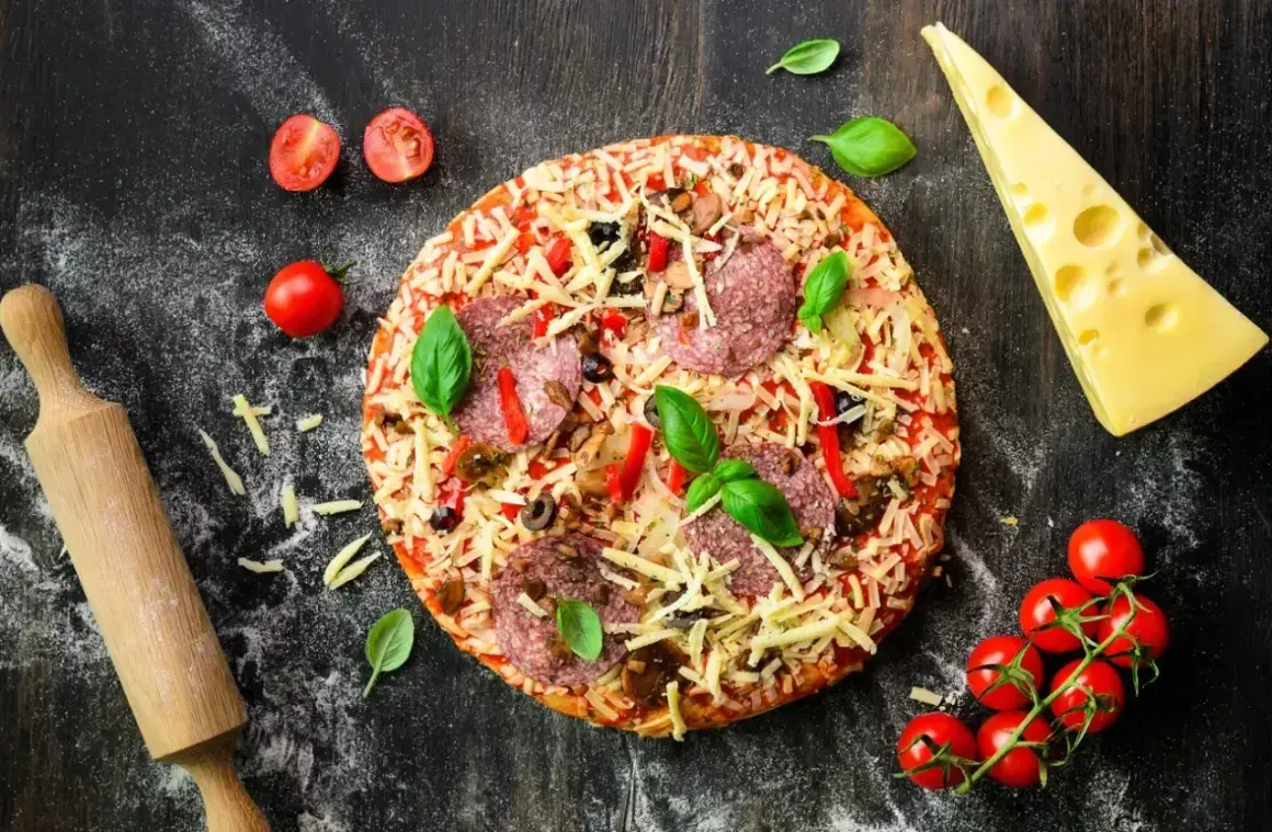 Gotowa pizza z salami i serem, obok wałek, pomidorki i ser. Idealna do domowego wypieku, zastanawiasz się, jaka mąka do pizzy domowej będzie najlepsza?