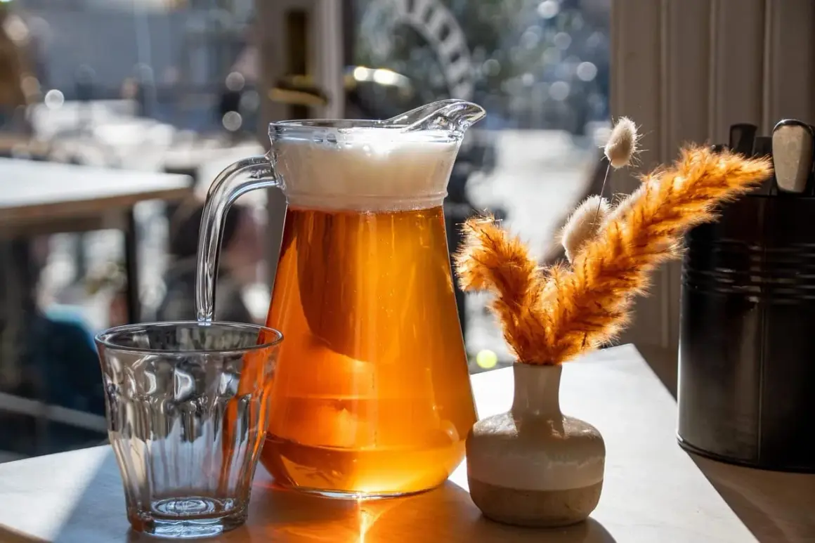 Dzbanek orzeźwiającego helles bier z pianką obok szklanki i ozdobnej trawy.