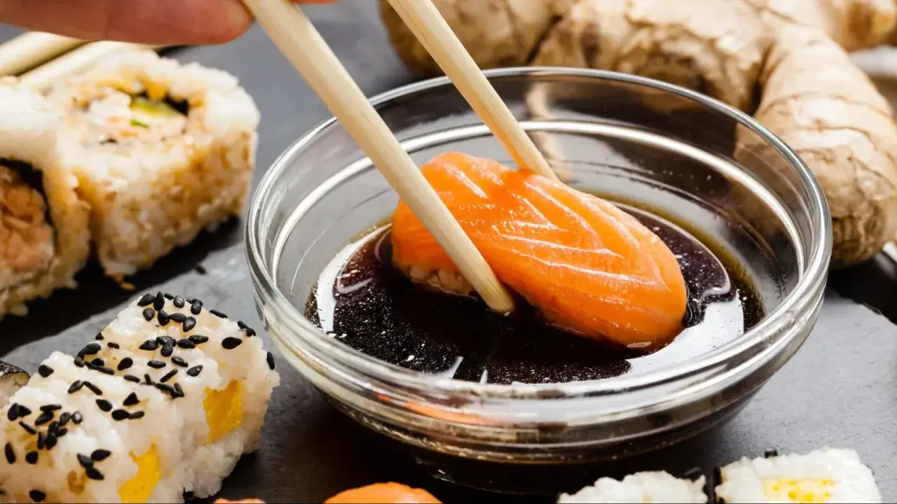 Chopsticks zanurzają kawałek łososia do sosu sojowego, przygotowując go do sushi. Obok leżą rolki sushi i imbir.