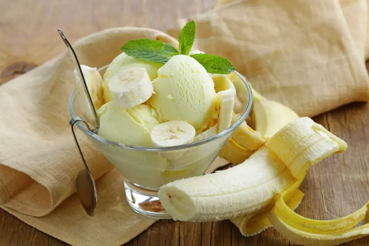 Pyszne lody bananowe w pucharku, ozdobione plasterkami banana i listkami mięty. Obok leży obrany banan.