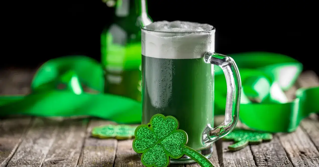Zielone piwo irlandzkie w kuflu, ozdobione koniczynką, na tle zielonej wstążki i butelki.