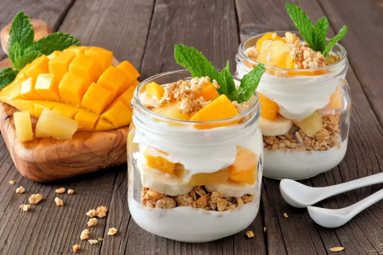 Dwa słoiki pysznych lodów jogurtowych z mango, bananem i granolą, udekorowane miętą. Obok pokrojone mango na drewnianej desce.