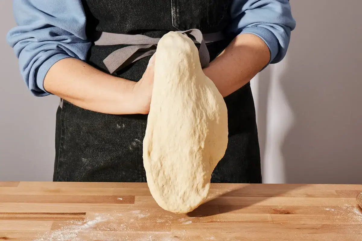 Ręce formują elastyczne ciasto na pizzę, które zwisa na drewnianym blacie.