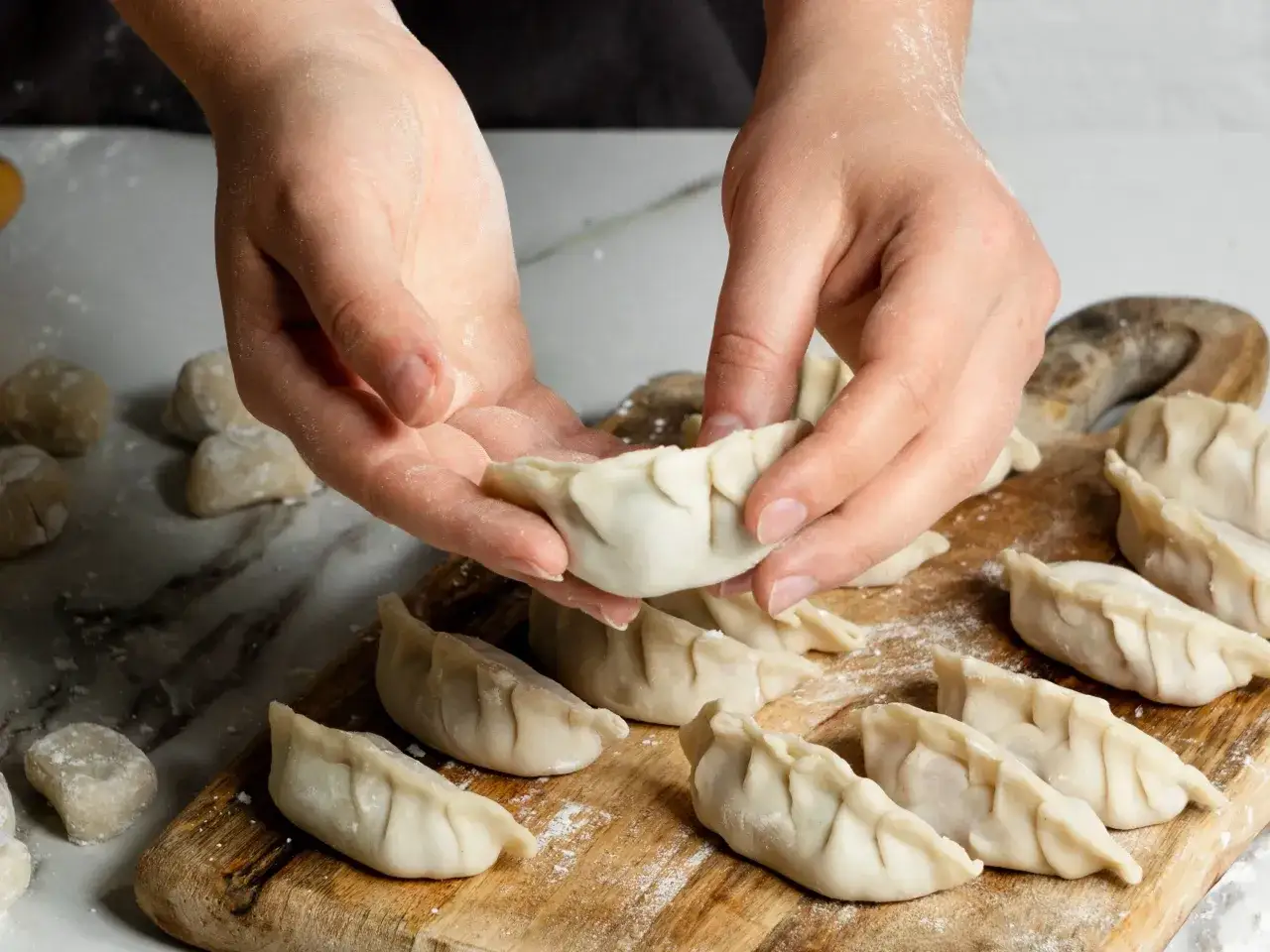 Ręce Ani formują pierogi na drewnianej desce. Widać gotowe pierożki i surowe ciasto.