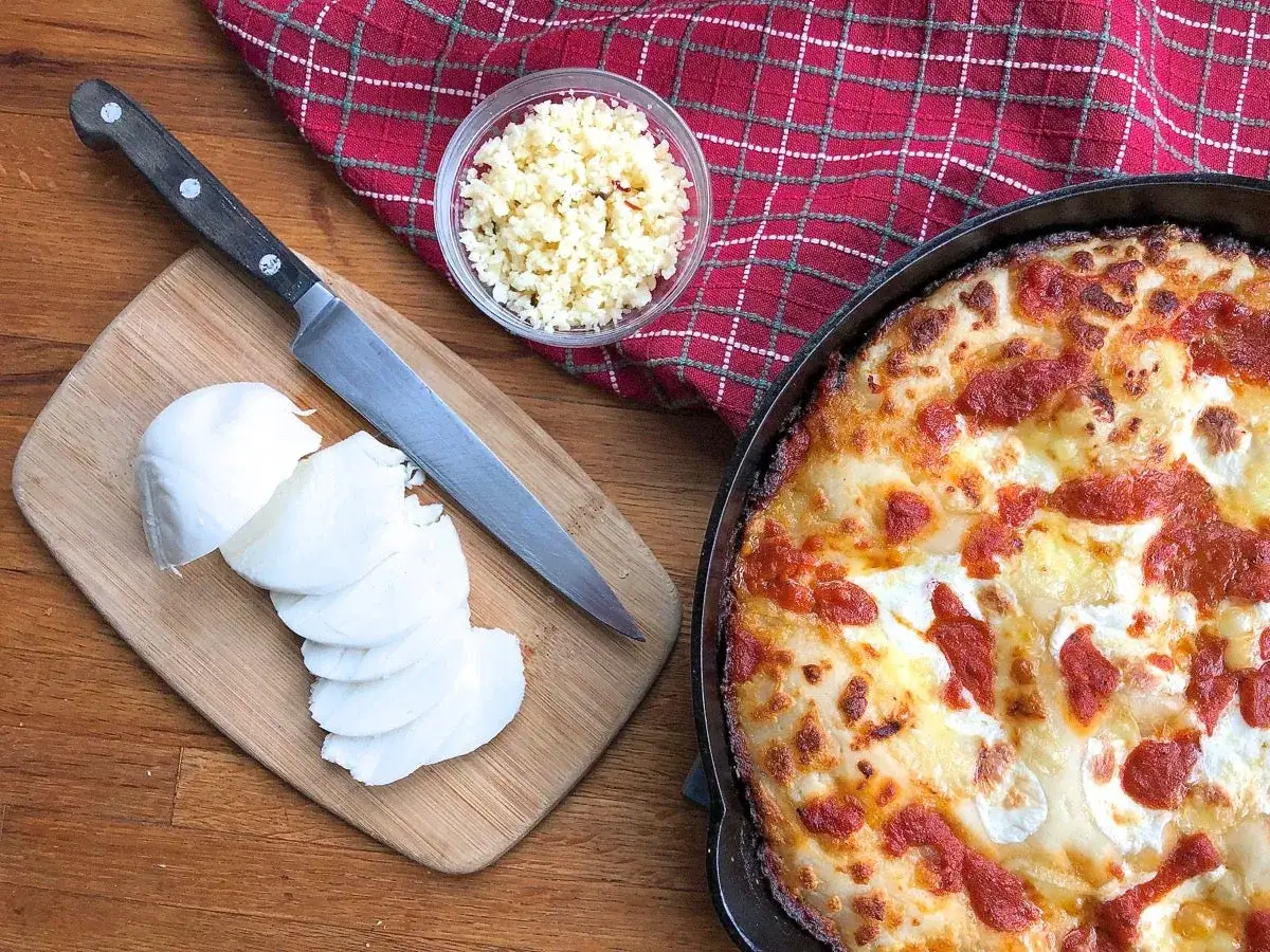 Idealny jaki ser do pizzy? Na desce pokrojona mozzarella, obok nóż i miseczka z tartym serem. W tle gorąca pizza.