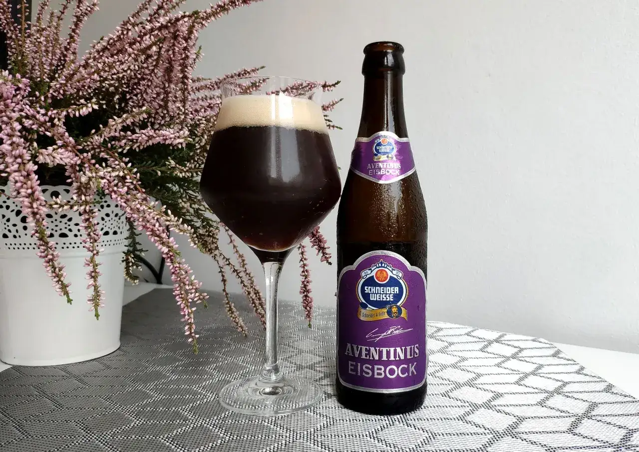 Schneider Weisse Aventinus Eisbock, ciemne piwo po procesie wymrażania, w szkle i butelce, obok kwitnącej wrzosy.