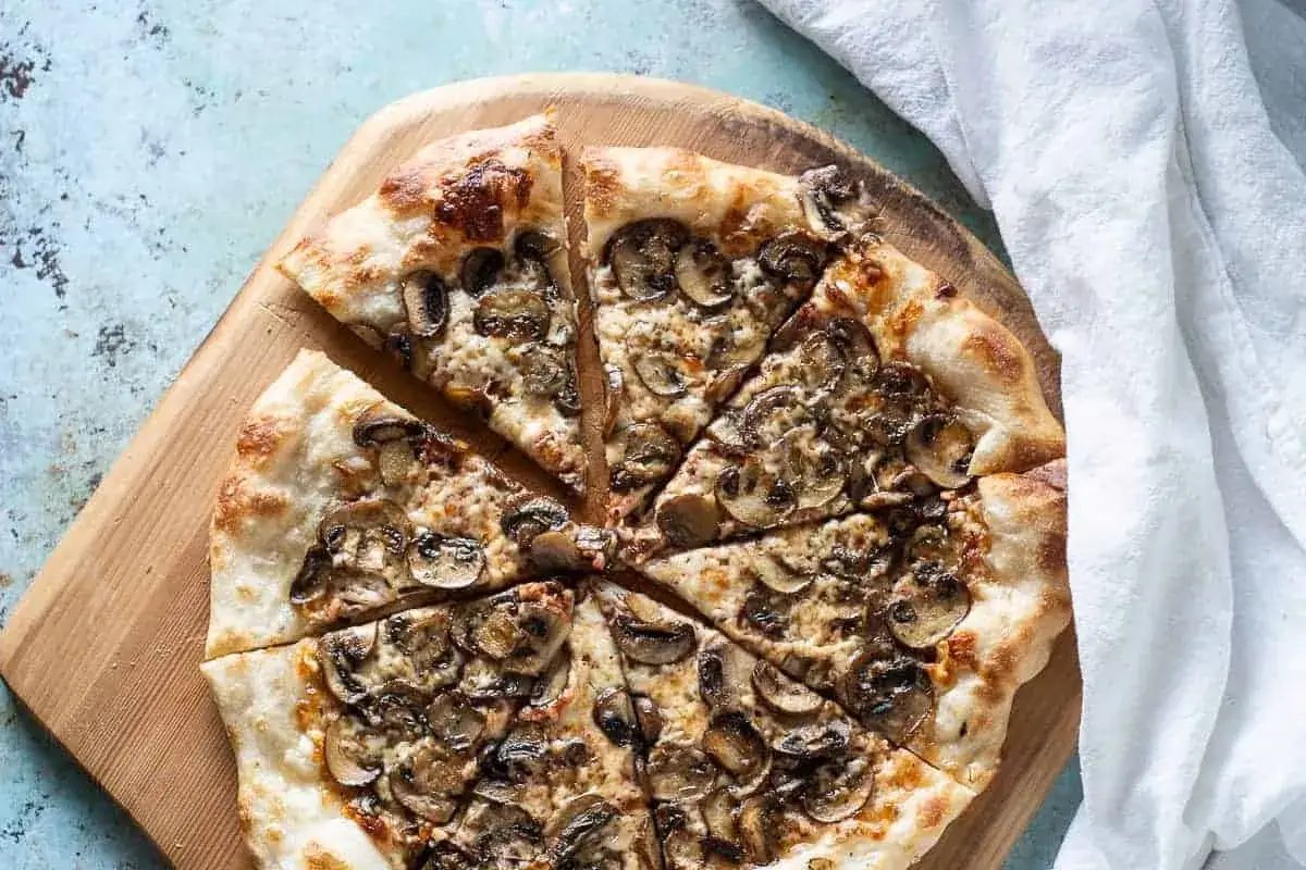 Pokrojona pizza z obfitym dodatkiem surowych pieczarek na pizze, podana na drewnianej desce.