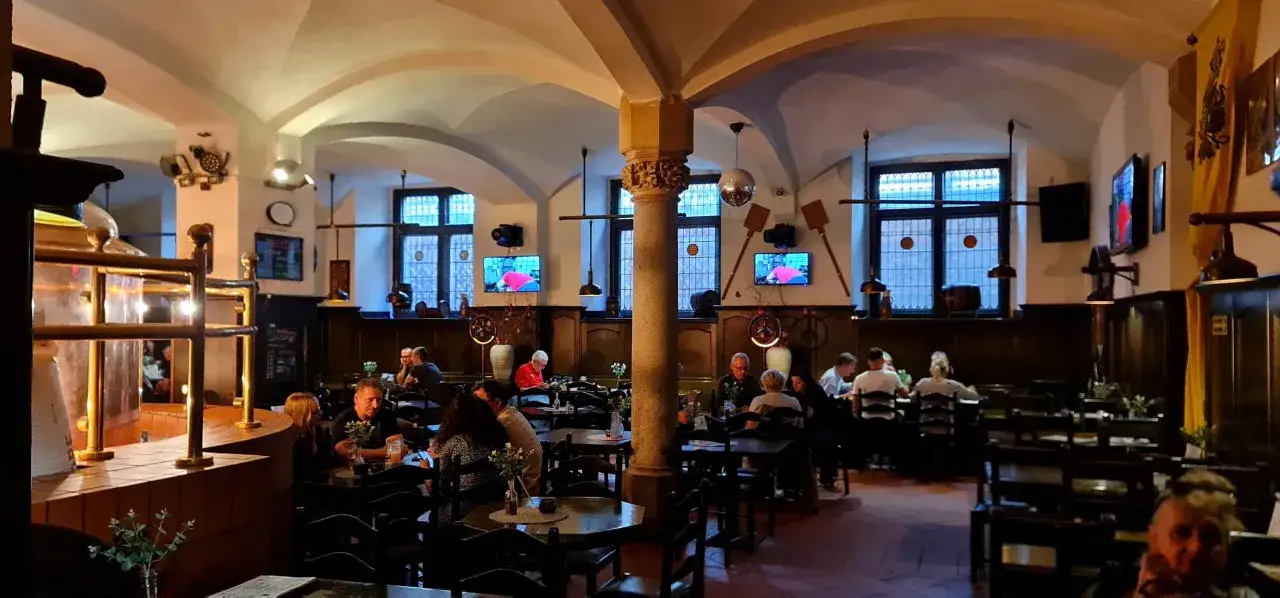 Wnętrze restauracji z widocznym kadłubem warzelni, gdzie serwują pyszne piwo spiż. Ludzie siedzą przy stołach, ciesząc się wieczorem.