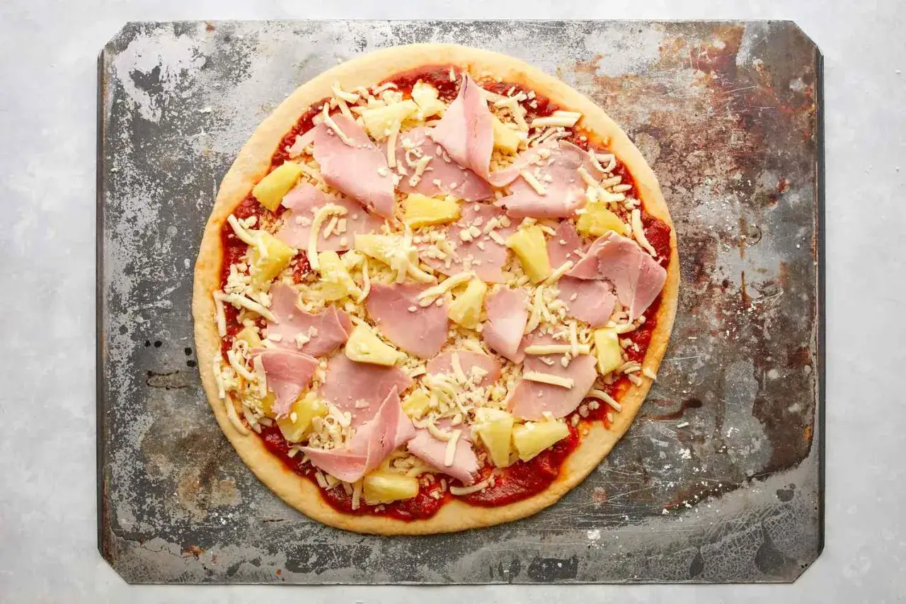 Gotowa do pieczenia pizza hawajska z szynką i ananasem, posypana serem, leży na metalowej blasze.
