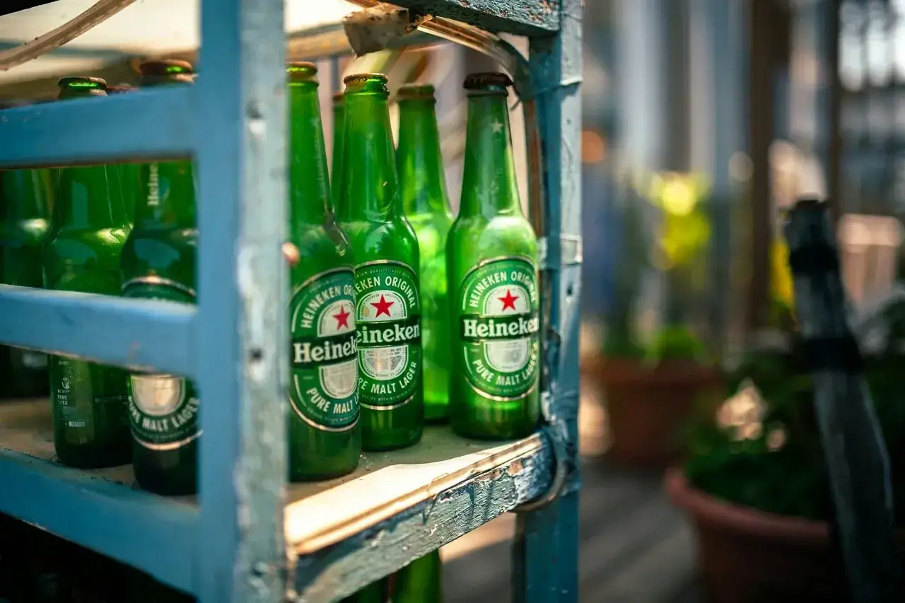 Zielone butelki piwa Heineken stoją na półce, w tle rośliny.