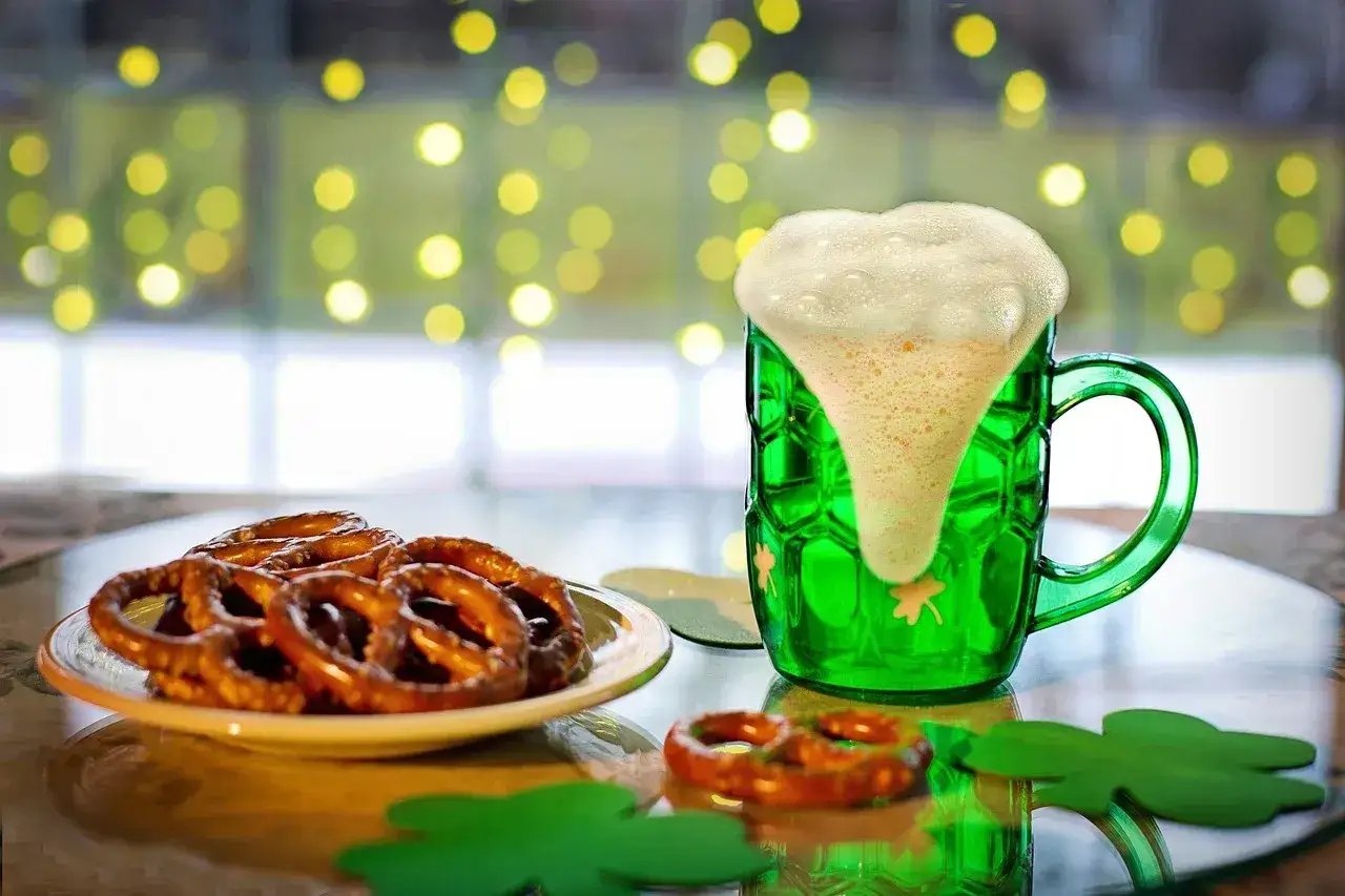Zielony kufel z obficie spienionym piwem stout, obok talerz precli i ozdoby w kształcie koniczyny.