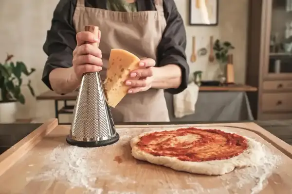 Ręce trzymają kawałek sera i tarkę, przygotowując się do starcia go na pizzę. To idealny sposób, czym posmarować pizzę, by była pyszna!