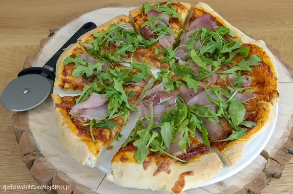 Pyszne pomysły na pizzę: cienkie ciasto, sos pomidorowy, ser, szynka i rukola. Obok leży nóż do pizzy.