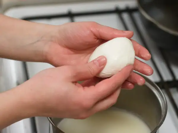 Dłonie trzymają kulkę mozzarelli nad garnkiem z mlekiem, sprawdzając, czy mozzarella w kulce się rozpuszcza.