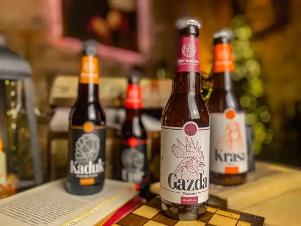 Piwo z Pomorza: butelki "Kaduk", "Gazda" i "Krasa" stoją na stole obok otwartej książki i szachownicy.