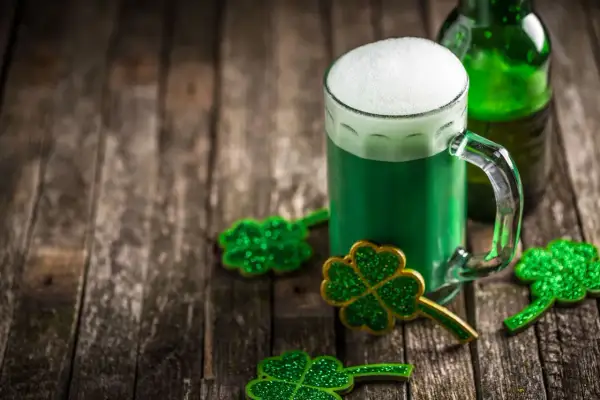 Zielone piwo irlandzkie z pianką, butelka i błyszczące koniczynki na drewnianym stole.