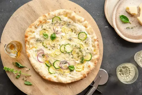Pyszna pizza bez sosu pomidorowego z cukinią, cebulą i ziołami, podana na drewnianej desce.