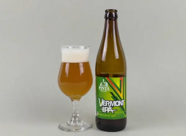 Szklanka z bursztynowym piwem Vermont IPA i butelka tego piwa. Idealne neipa piwo na wieczór.