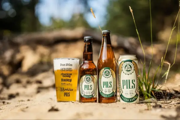 Szklanka, butelka i puszka piwa lager "Pils" od Trzech Kumpli na piasku, wśród traw.
