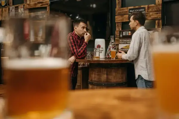 Dwóch mężczyzn rozmawia przy barze, zastanawiając się, ile węglowodanów ma piwo. Na stole stoją kufle i beczka.