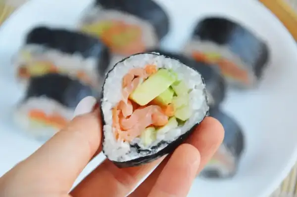 Ręka trzyma kawałek sushi z łososiem i awokado w środku. Na talerzu widać więcej takich roladek.