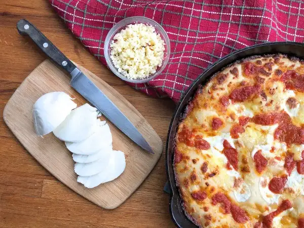 Idealny jaki ser do pizzy? Na desce świeża mozzarella, obok nóż i miseczka z tartym serem. Obok pyszna, gorąca pizza.