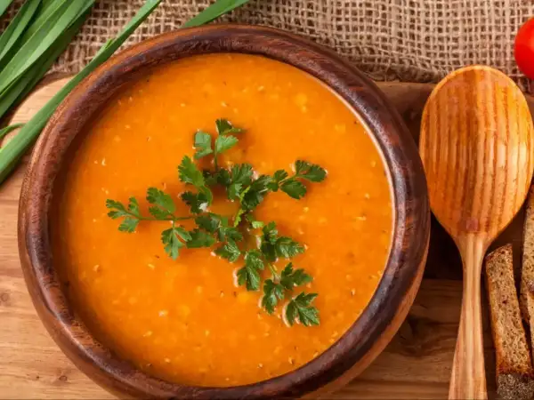 Aksamitna zupa pomidorowa w drewnianej misce, udekorowana natką pietruszki. Obok łyżka i kromki chleba.