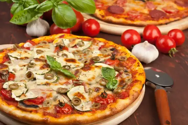 Pyszna pizza z cukinią, oliwkami i pomidorkami, obok surowe pieczarki na pizze, czosnek i bazylia.