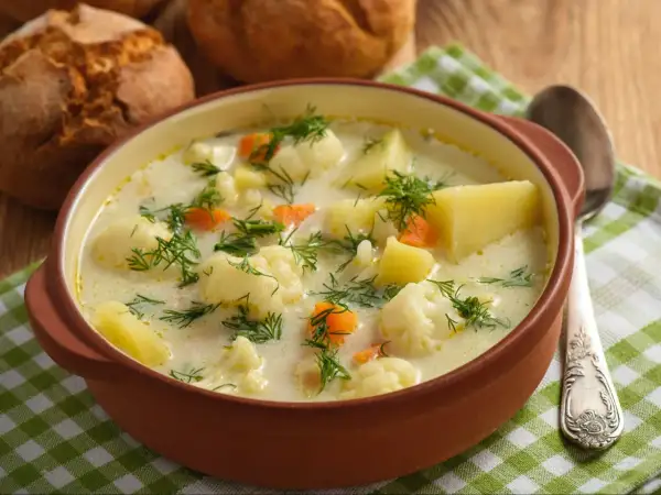 Aksamitna zupa kalafiorowa jak u babci, z ziemniakami i marchewką, posypana koperkiem. Obok bułeczki i łyżka.